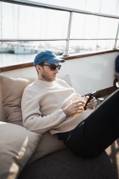 Young man enjoying leisure time on a yacht with a smartphone and drink in hand.