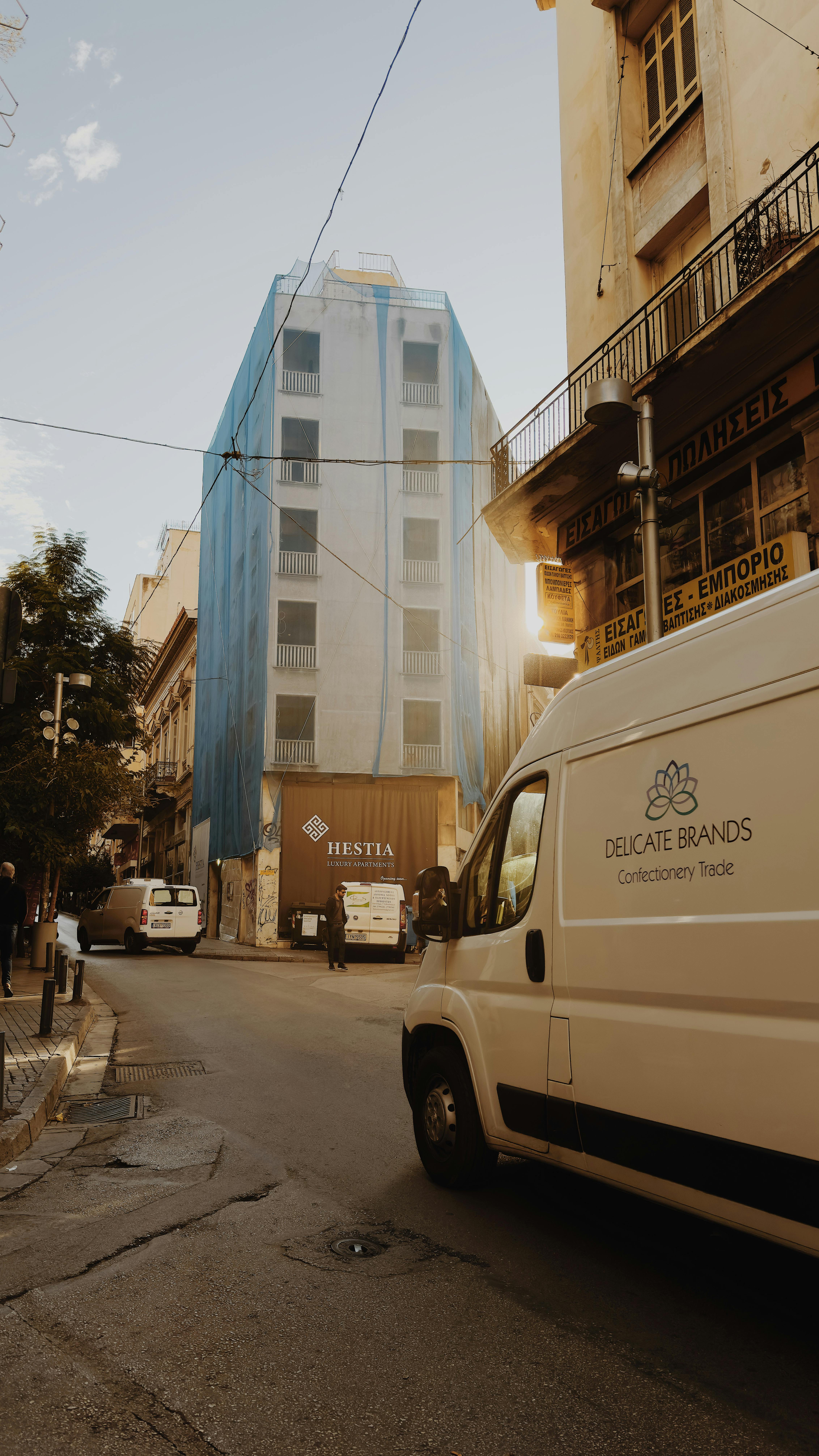 Free Urban street view featuring modern building facade with parked vans and pedestrians in warm lighting. Stock Photo