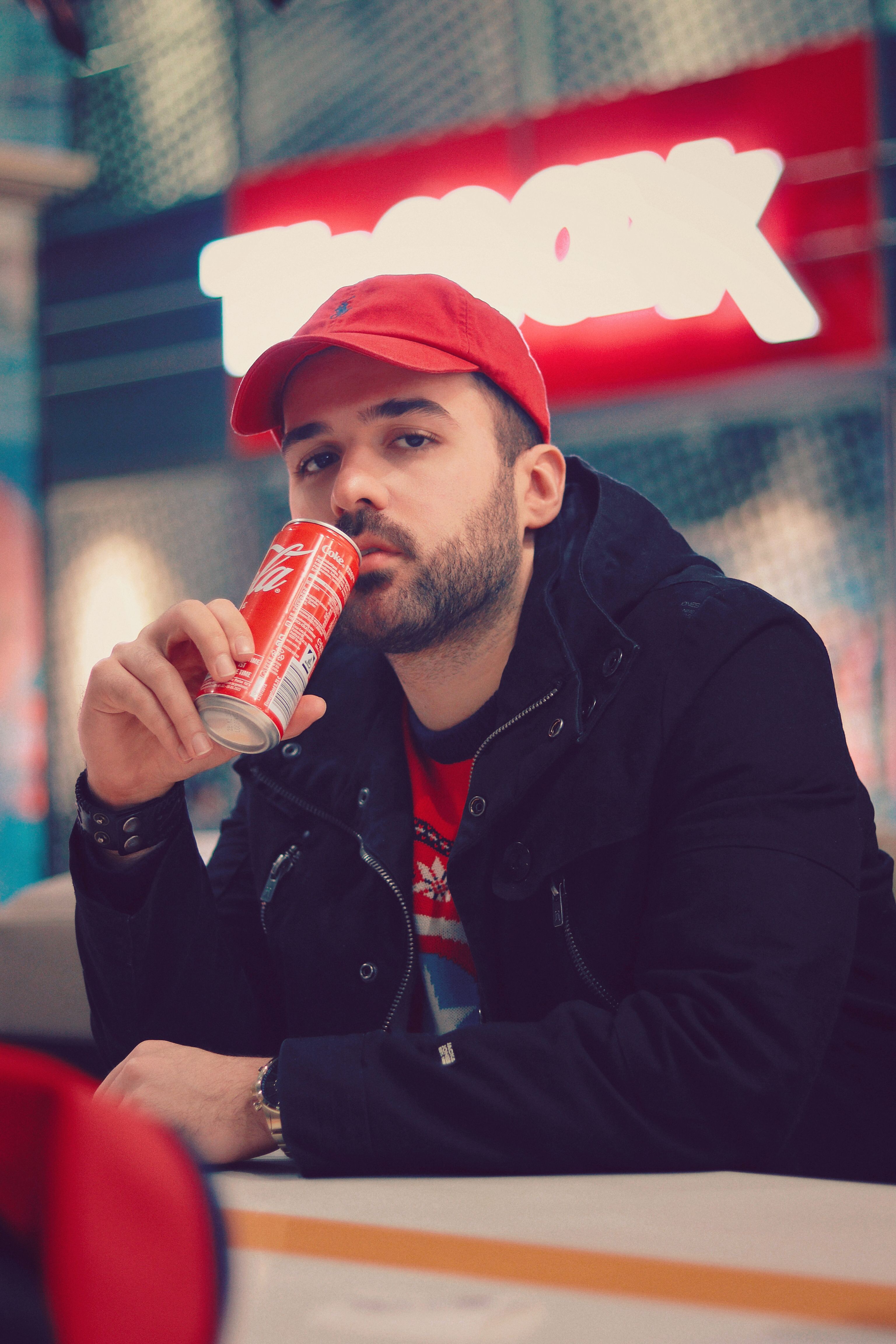 Man in red cap drinking soda indoors, with vibrant urban background.