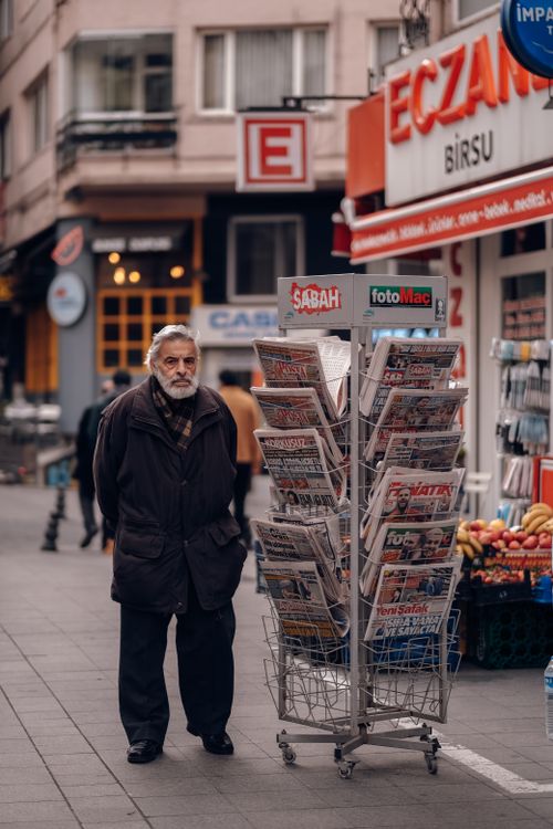 Free Elderly man stands by a newspaper stand on a bustling street with shops in Turkey. Stock Photo