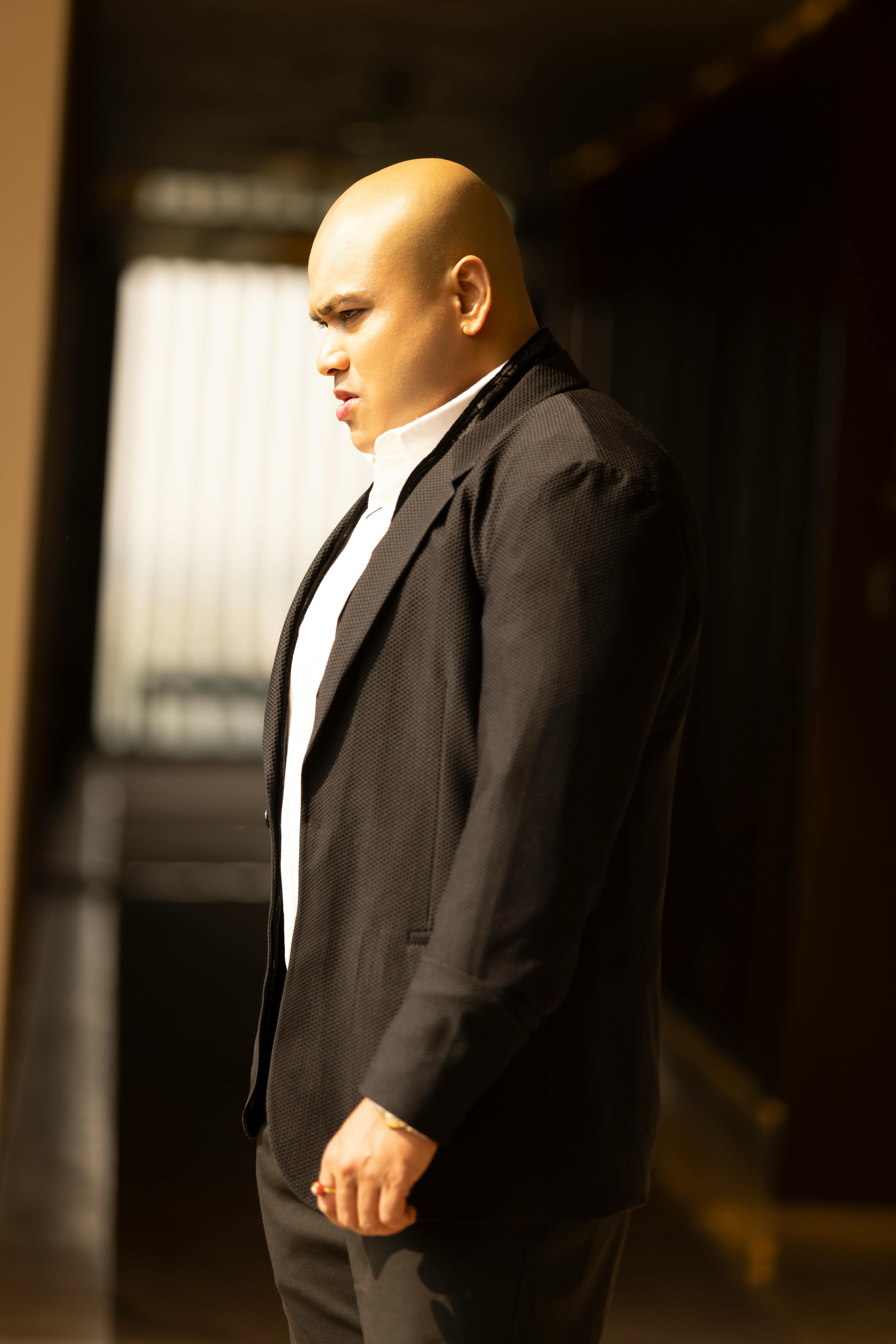 A well-dressed man in a black suit stands indoors with soft natural lighting.