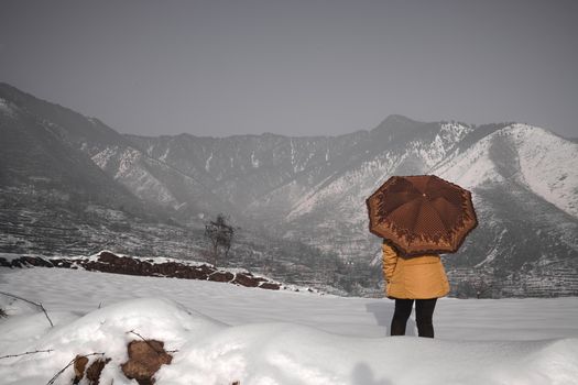 A person in winter attire holding an umbrella, standing on snow-covered ground with mountain scenery.