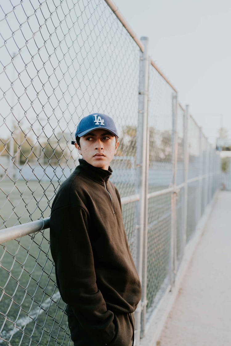 Man In A Sweatshirt And A Cap By A Chain Link Fence