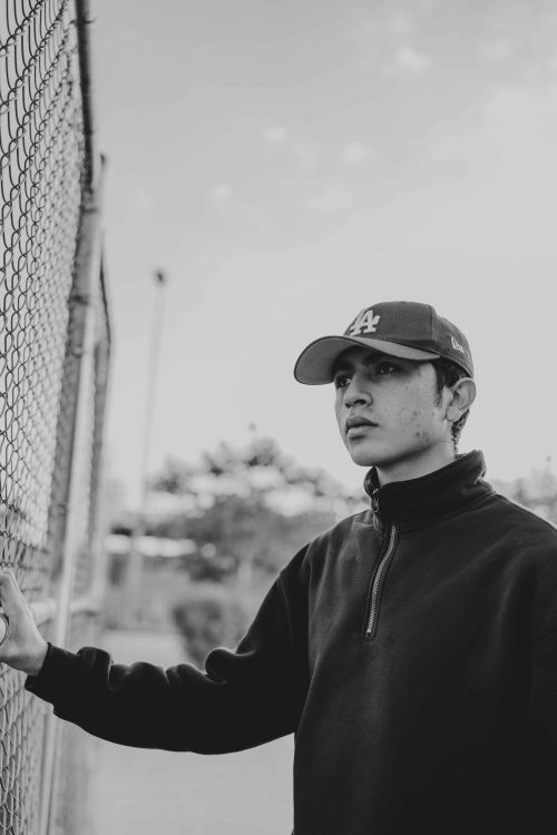 Free A black and white portrait of a teenage boy standing by a fence wearing a cap. Stock Photo