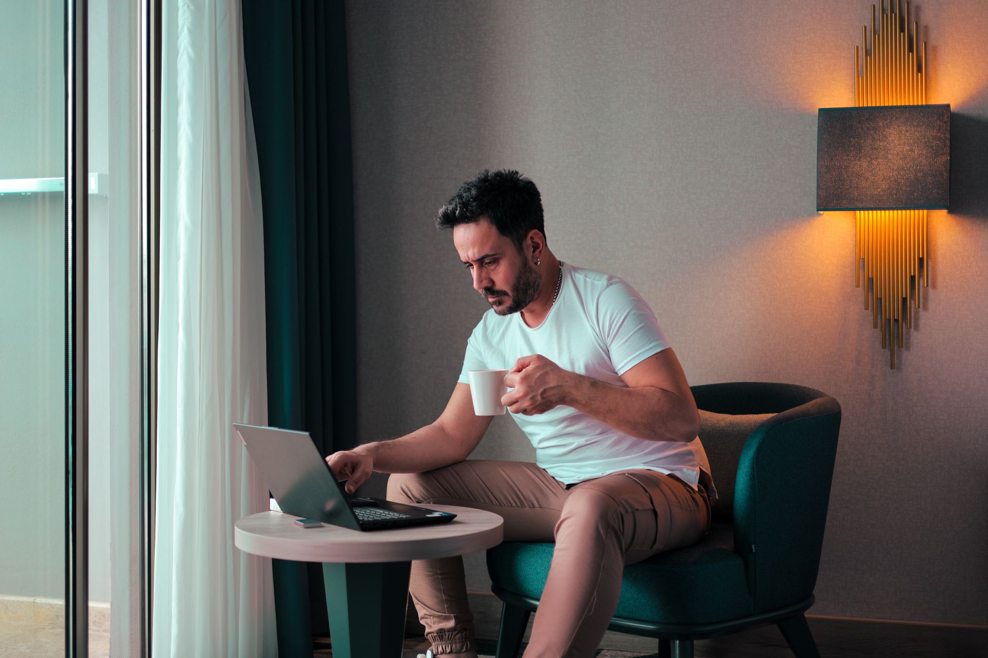 A traveler working on a laptop in a hotel room, holding a cup of coffee