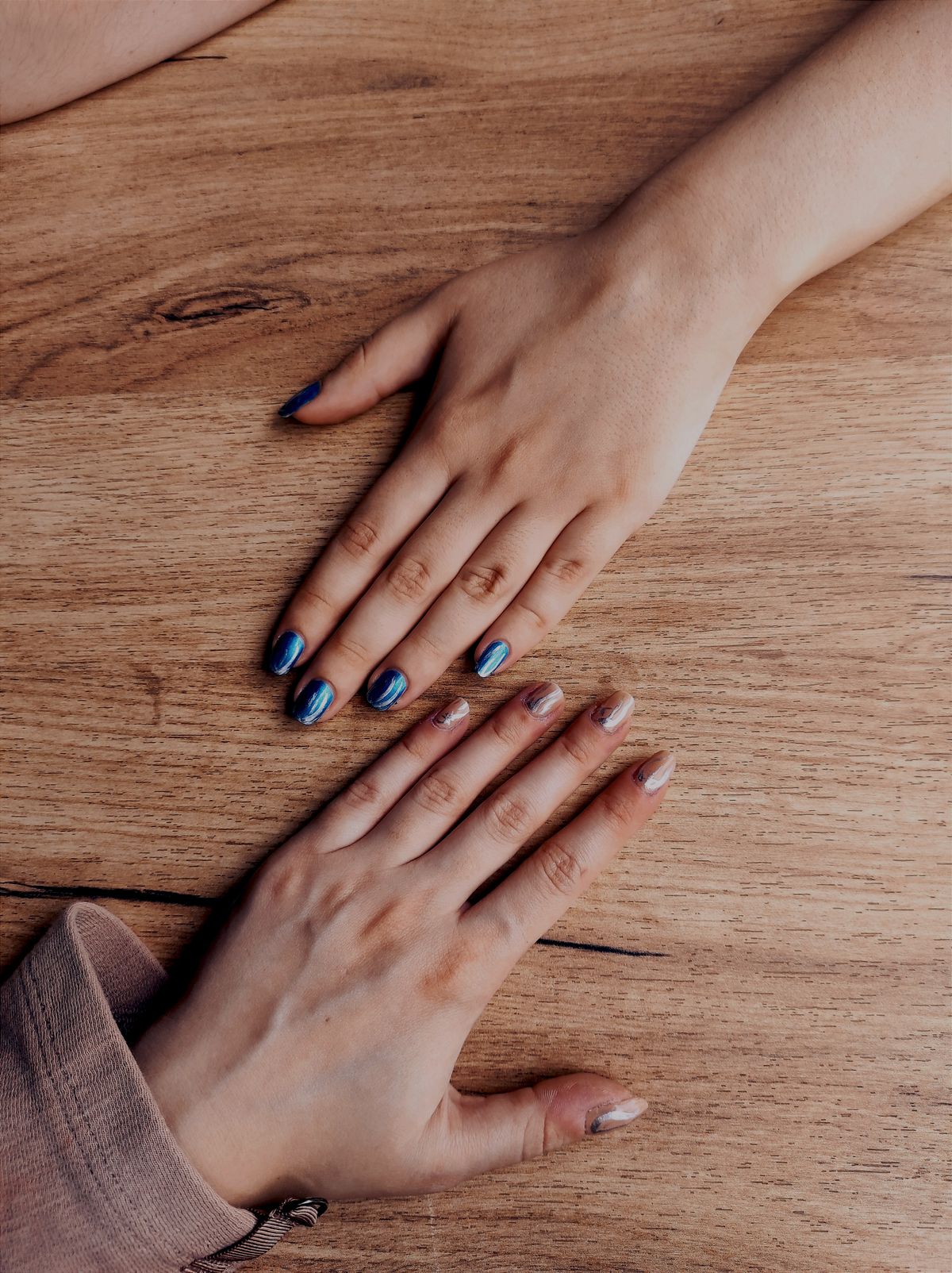 Two sets of hands with painted nails laid on a wooden table, showcasing diverse nail art designs.
