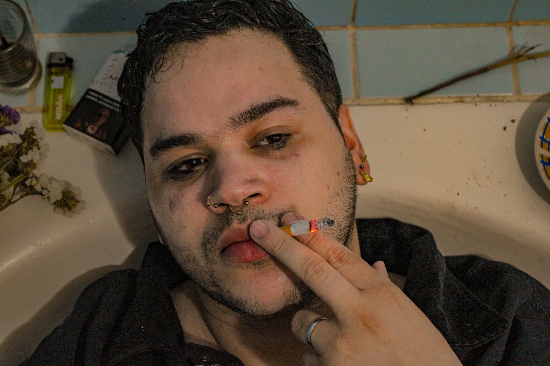 A troubled man with piercings smokes a cigarette, lying in a bathtub, reflecting deep thoughts.
