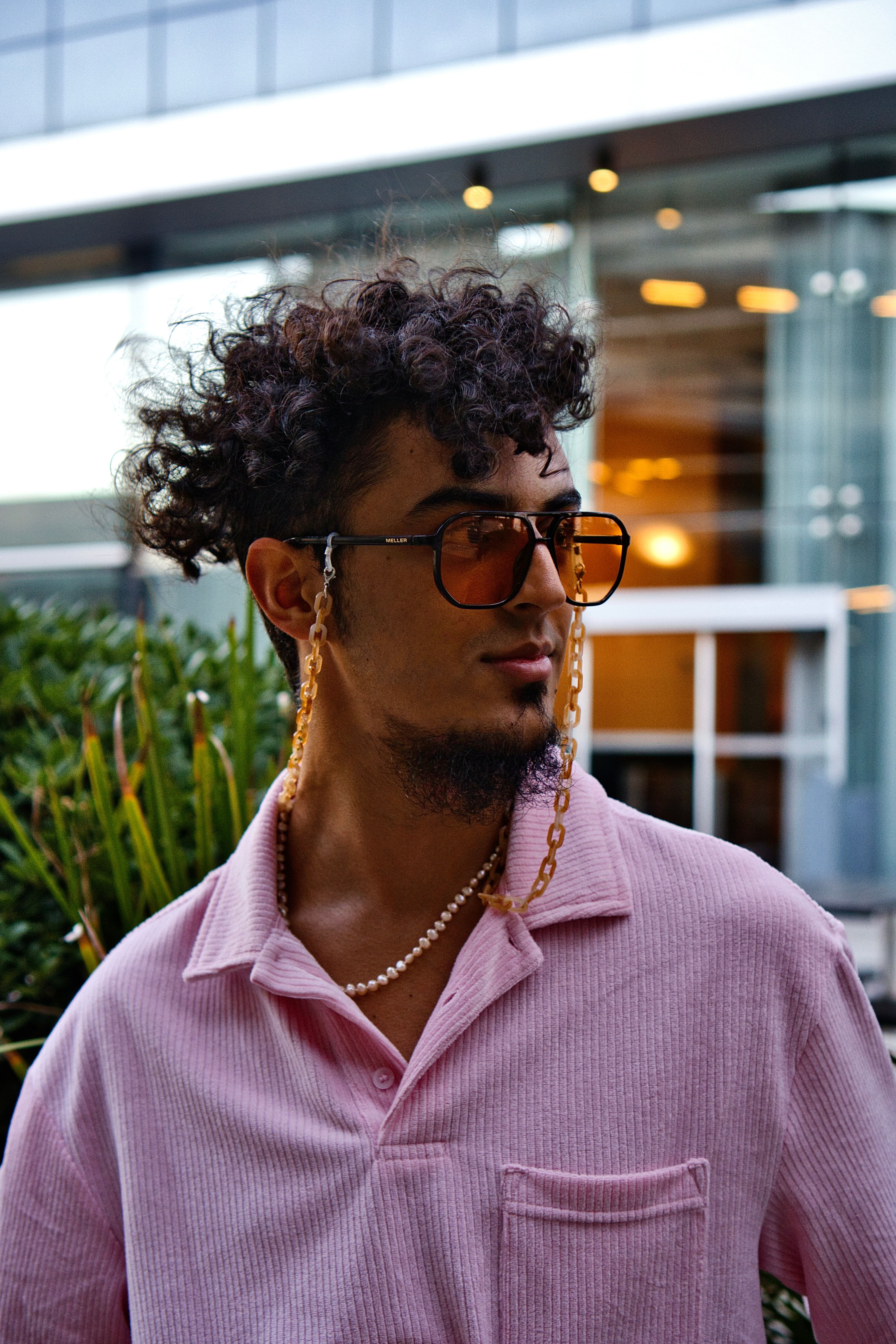 Trendy young man with curly hair and sunglasses, wearing a pink shirt, outdoor urban setting.