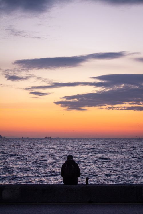 Free Silhouette of a person sitting by the sea, admiring a vibrant sunset. Stock Photo