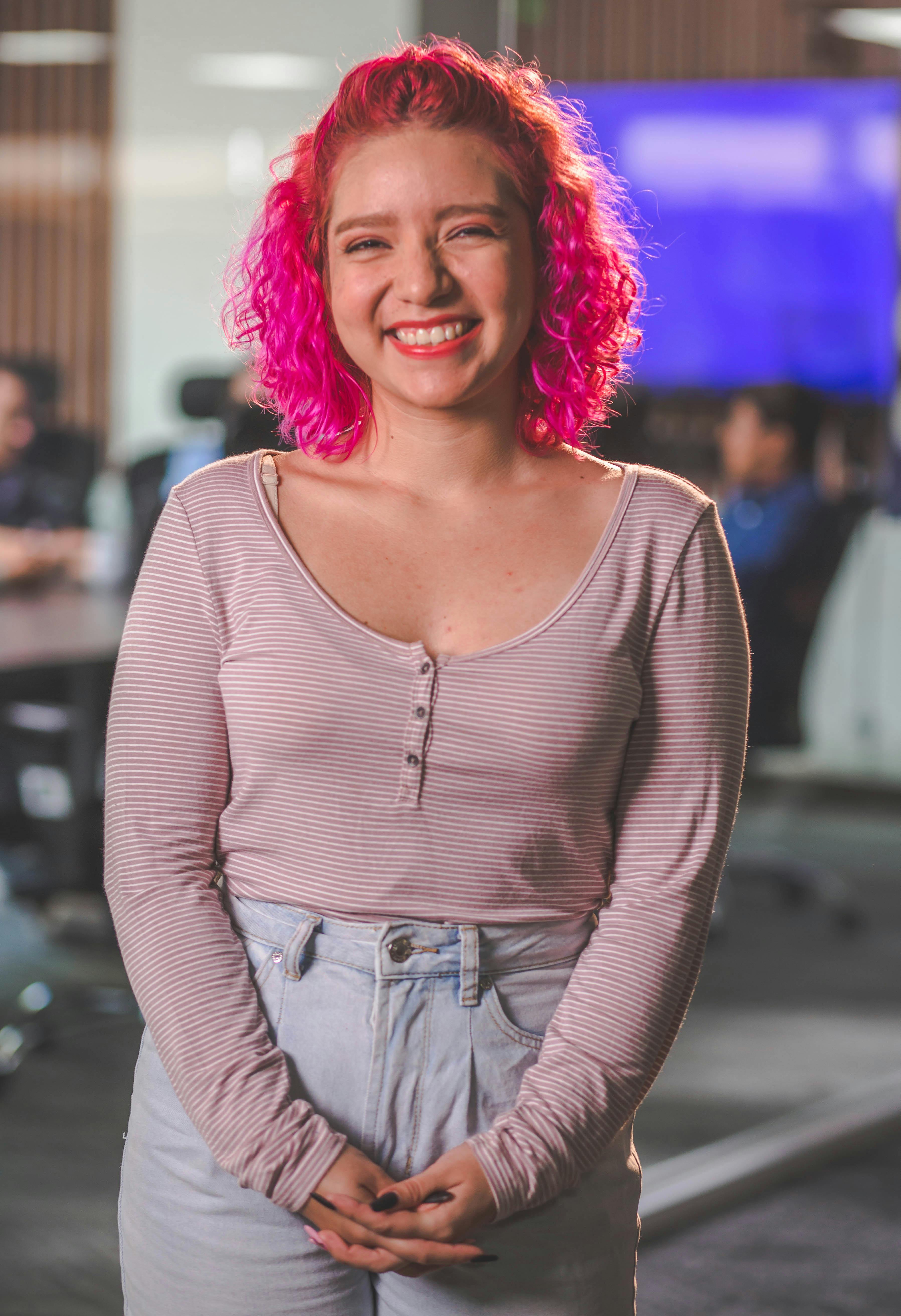 Confident woman with vibrant pink hair smiling in a casual office environment.