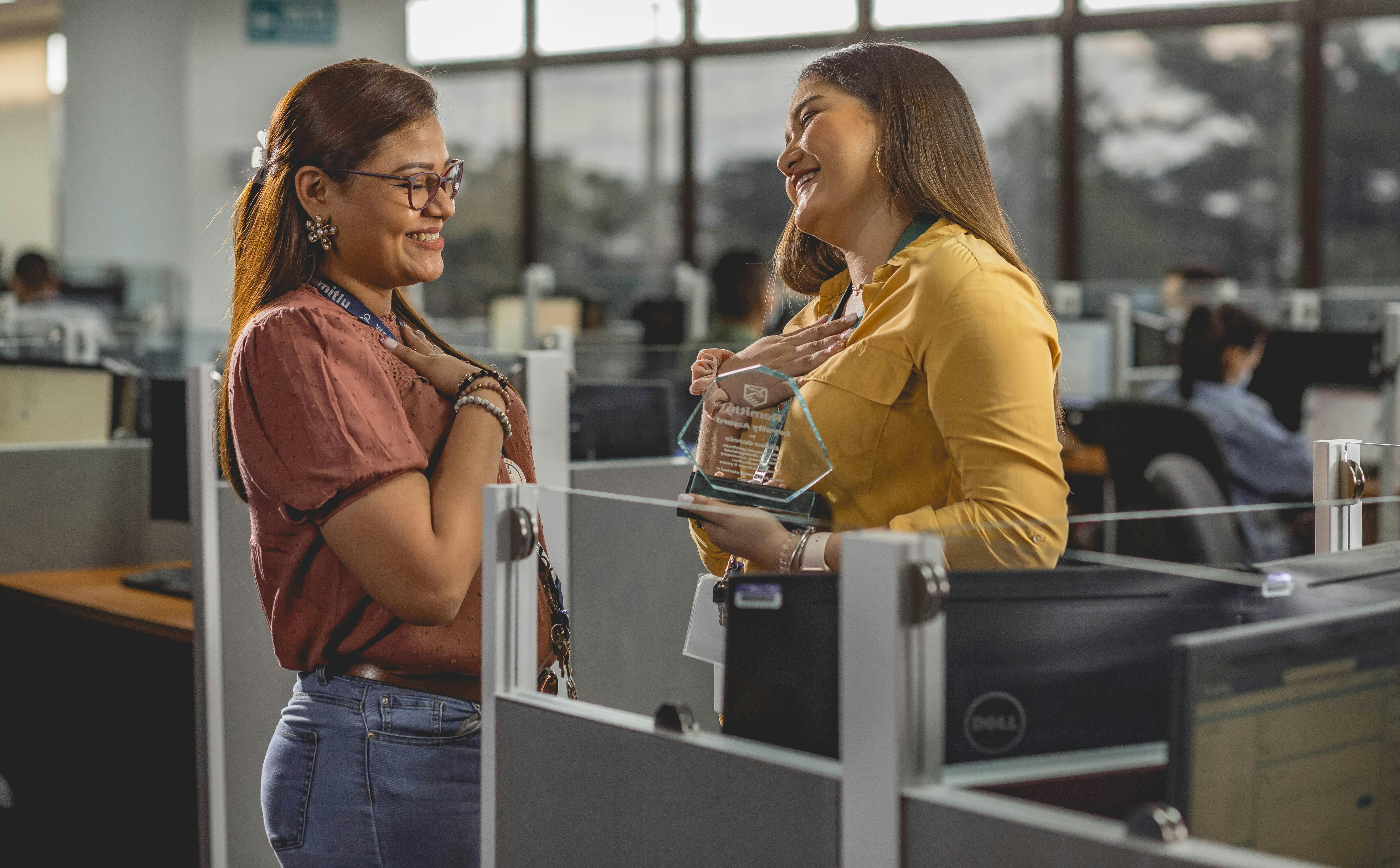 Two women smile and share a joyful moment receiving an award in a modern office setting.