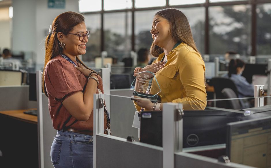 Duas mulheres sorriem e compartilham um momento alegre recebendo um prêmio em um ambiente de escritório moderno.
