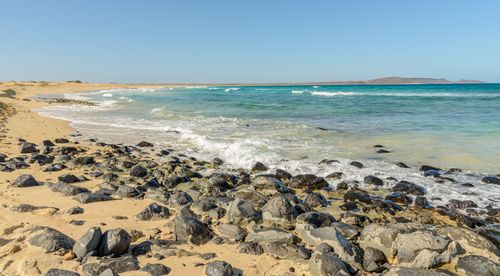 Free Tranquil beach scene with clear blue waters and rocky shore under a sunny sky. Stock Photo
