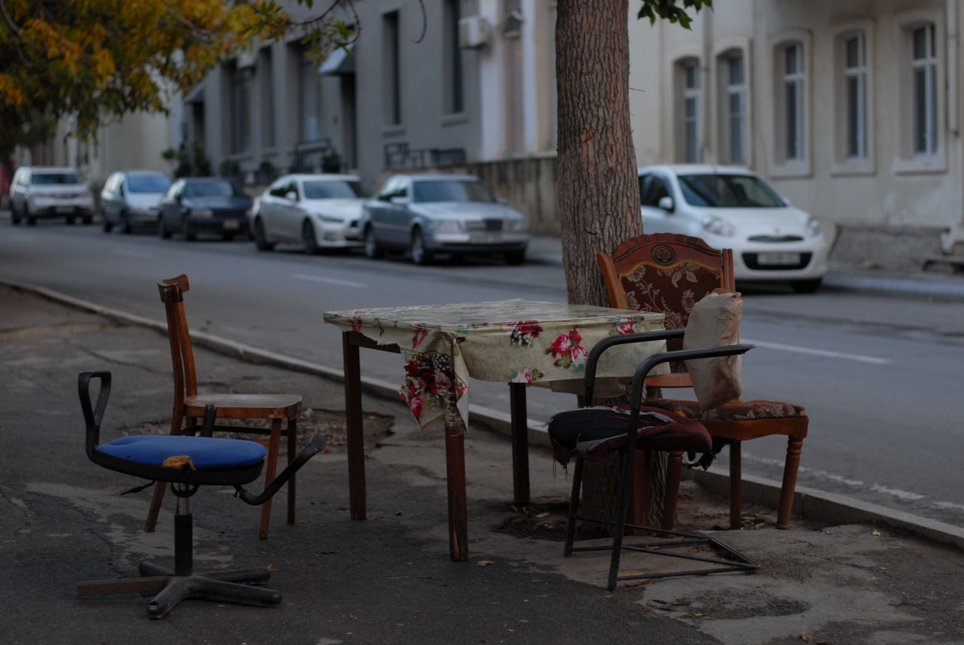 Mismatched chairs and table setup on a street sidewalk, surrounded by parked cars.