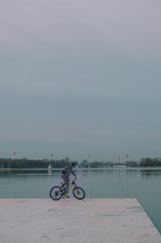 A person standing with a bicycle on a dock, gazing over a calm river under a serene sky.