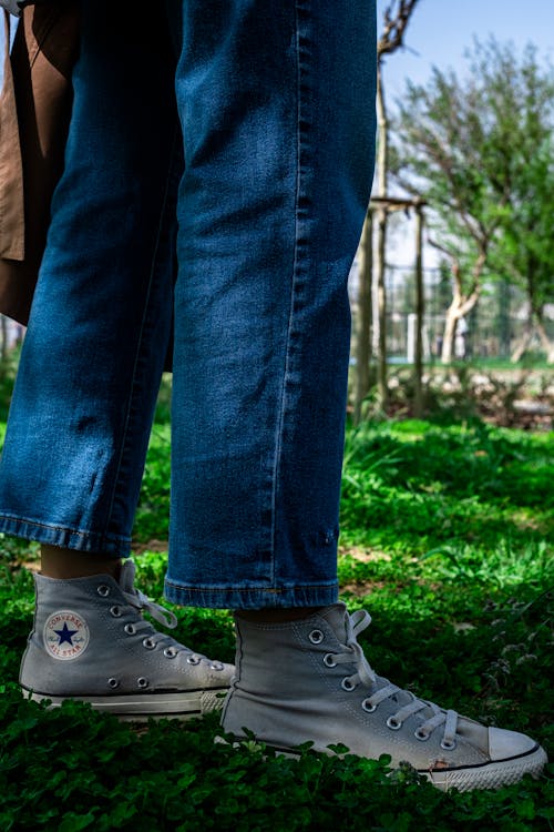 A Person in Denim Jeans Wearing Gray Converse Shoes while Standing on Grassy Ground