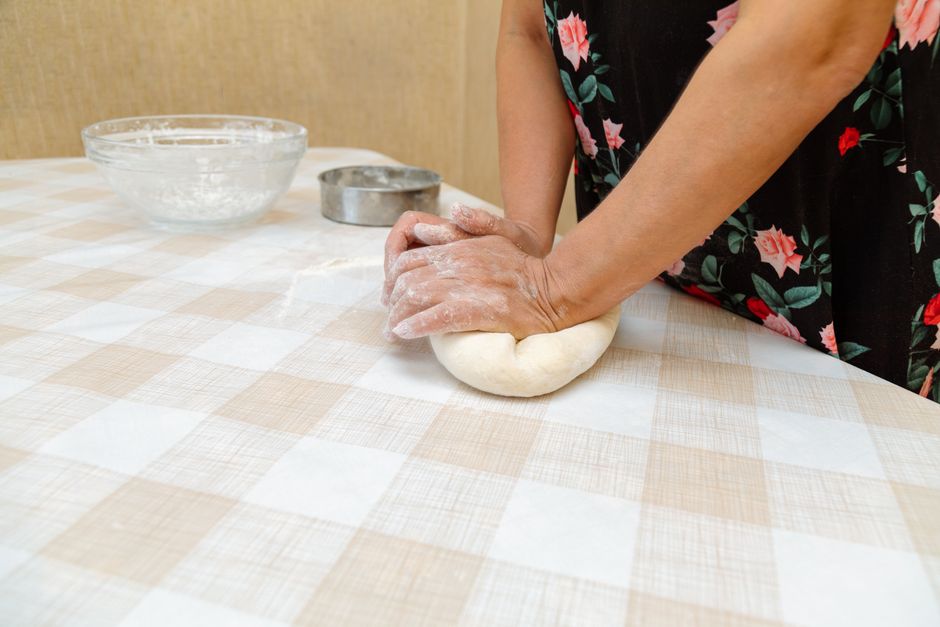 Hands of a woman kneading dough in a kitchen, floral dress visible.