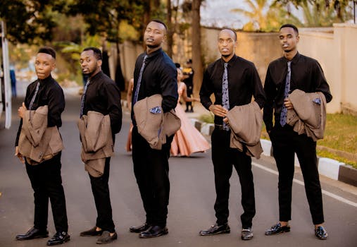 Five stylish men pose in formal attire outdoors, holding jackets on a street.