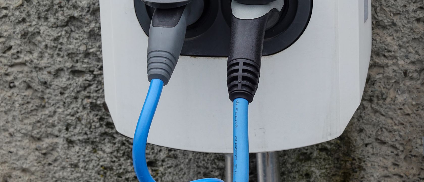 Close-up of an electric vehicle charging station with two cables in sockets against a textured wall.