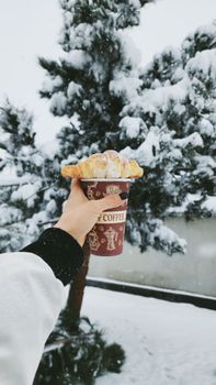 A hand holds a coffee cup with a croissant against a snowy pine tree background.