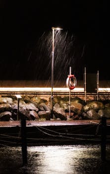 A dramatic street lamp illuminating rain over rocks by the shore at night.