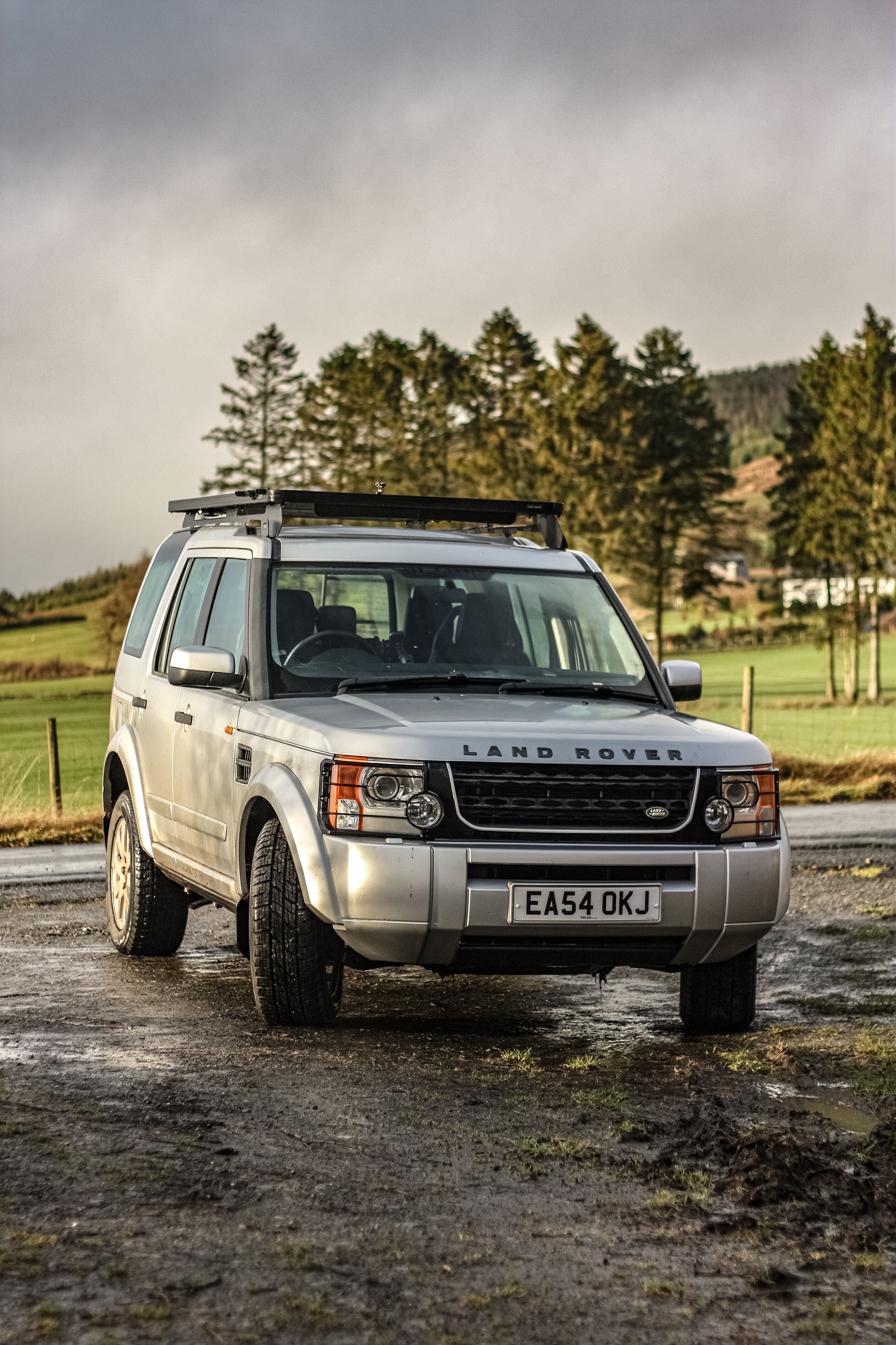 Land Rover parked on a muddy rural road with trees in the background.