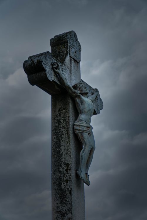 Free A detailed sculpture of Jesus on a cross under a dramatic cloudy sky, highlighting religious symbolism. Stock Photo