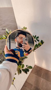 Man taking a selfie with a smartphone using a circular mirror reflecting his image indoors.