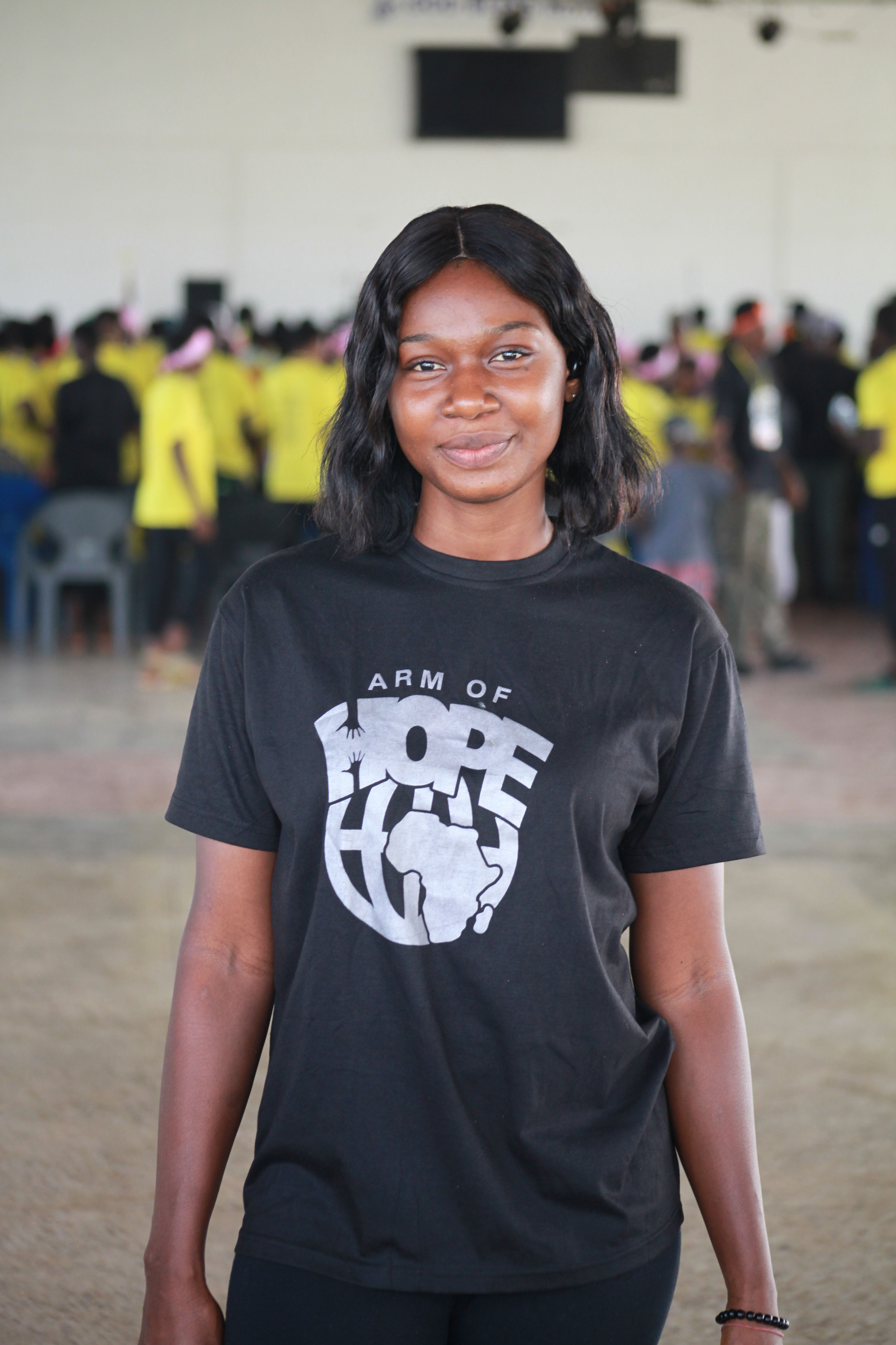 Smiling young woman wearing a black 'Arm of Hope' T-shirt at an indoor gathering.