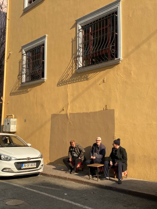 Free Three men sitting on stools by a sunlit yellow building's wall, with a parked car nearby. Stock Photo