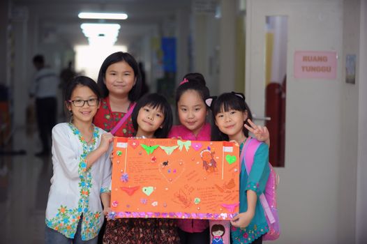 Five Asian schoolgirls joyfully presenting colorful artwork in a bright school corridor.