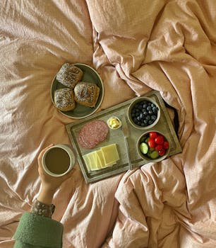 Top view of a cozy breakfast in bed with pastries, coffee, and fresh fruit on a tray.