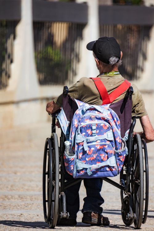 Free Disabled man in a wheelchair with a colorful backpack outdoors, wearing a baseball cap and casual attire. Stock Photo