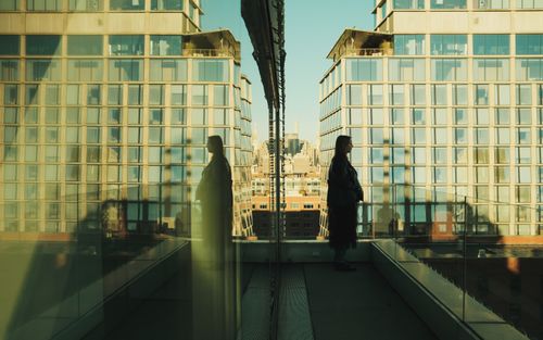 Free A woman stands on a New York balcony with reflections on a glass facade, showcasing an urban skyline. Stock Photo