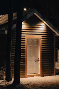 A warmly lit cabin with a white door surrounded by snow, creating a cozy winter night atmosphere.