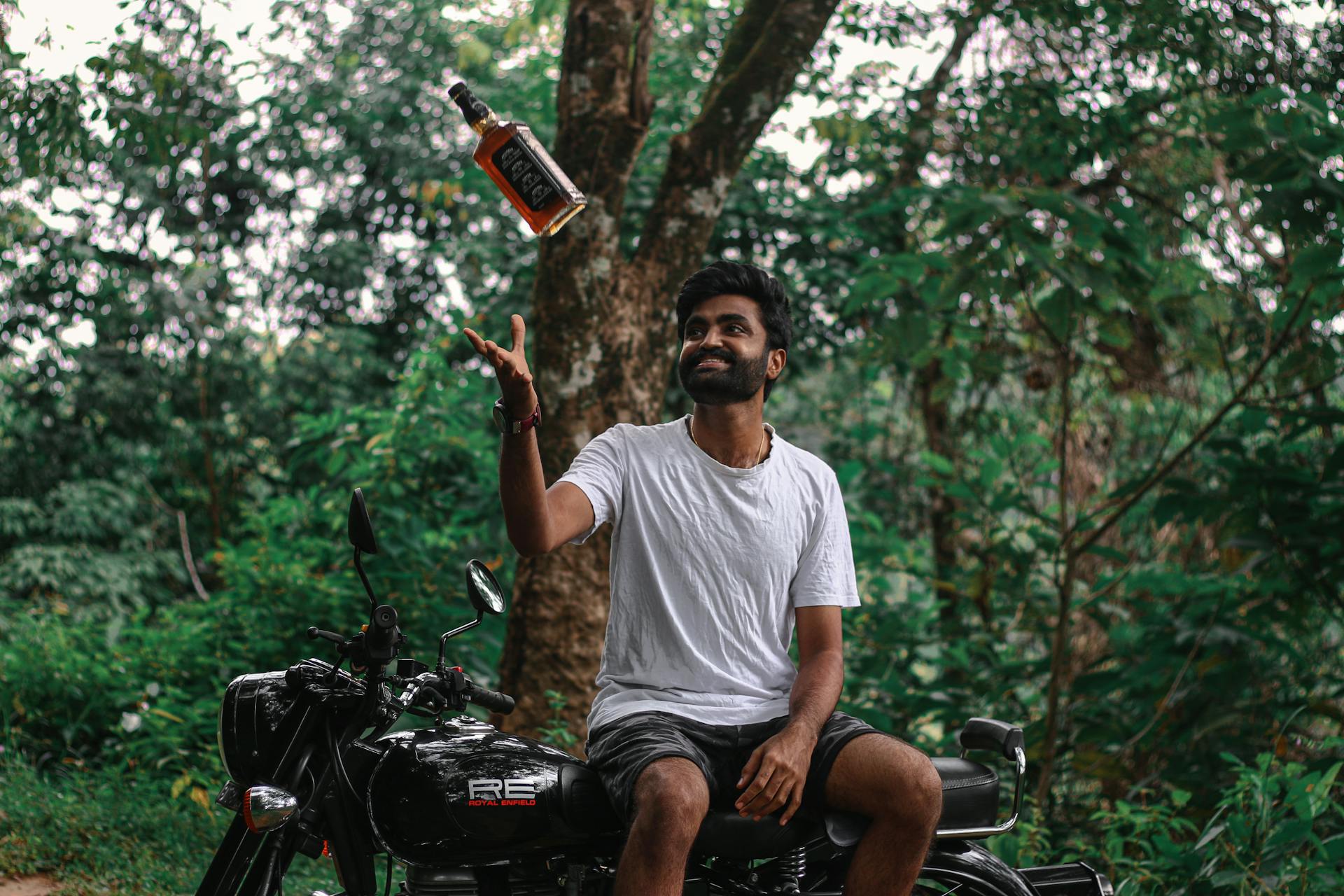 Smiling man sits on motorcycle, playfully tossing whiskey bottle in lush forest setting.