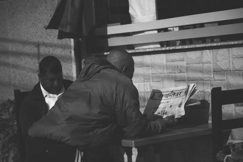 Grayscale Photo of Man Leaning on Table While Reading Newspaper