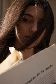 Close-up portrait of a young woman reading a fashion magazine with sunlight on her face.