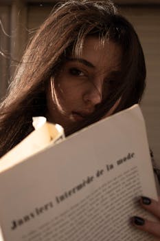 A young woman reads a fashion magazine indoors, bathed in warm light.
