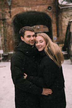 A young couple in winter coats embrace and smile as snowflakes fall around them.