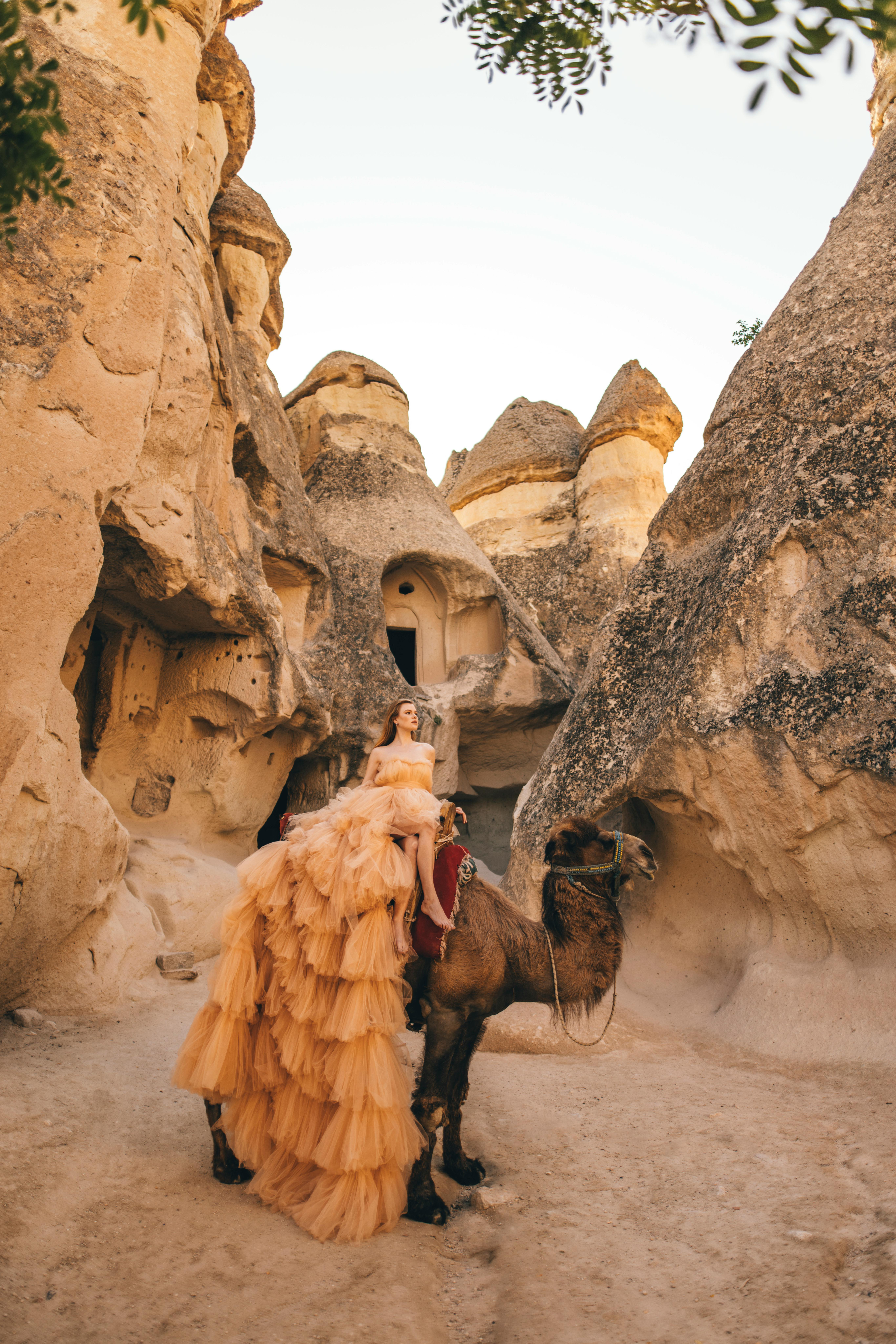 Woman in a flowing gown sits on a camel amidst the rocky formations of Cappadocia.