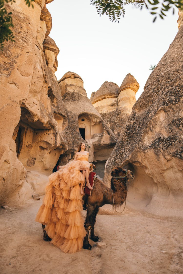 Woman In Gown Sitting On Camel Near Stone Caves