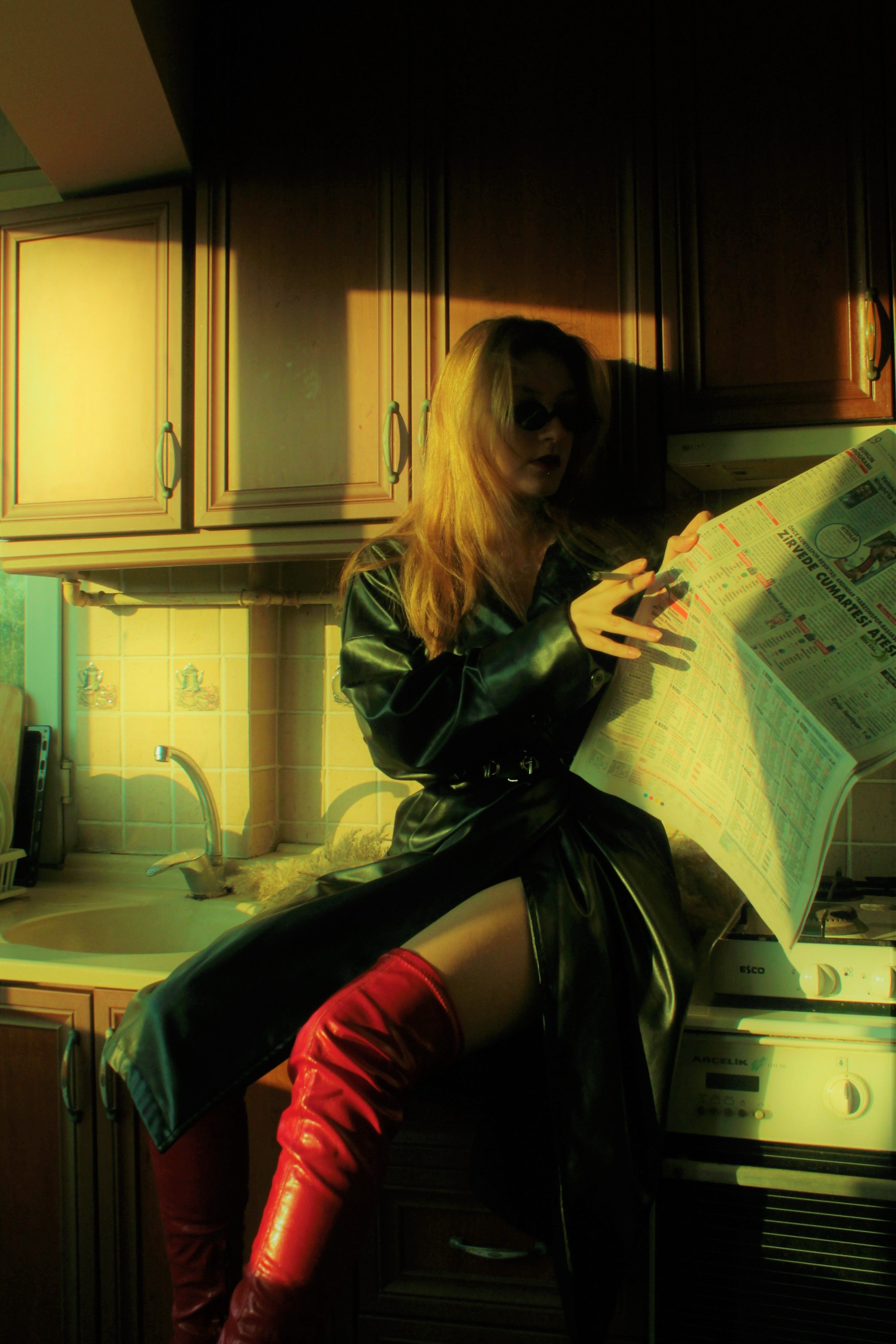 Elegant woman in sunglasses sitting in kitchen reading a newspaper, exuding sophistication and style.