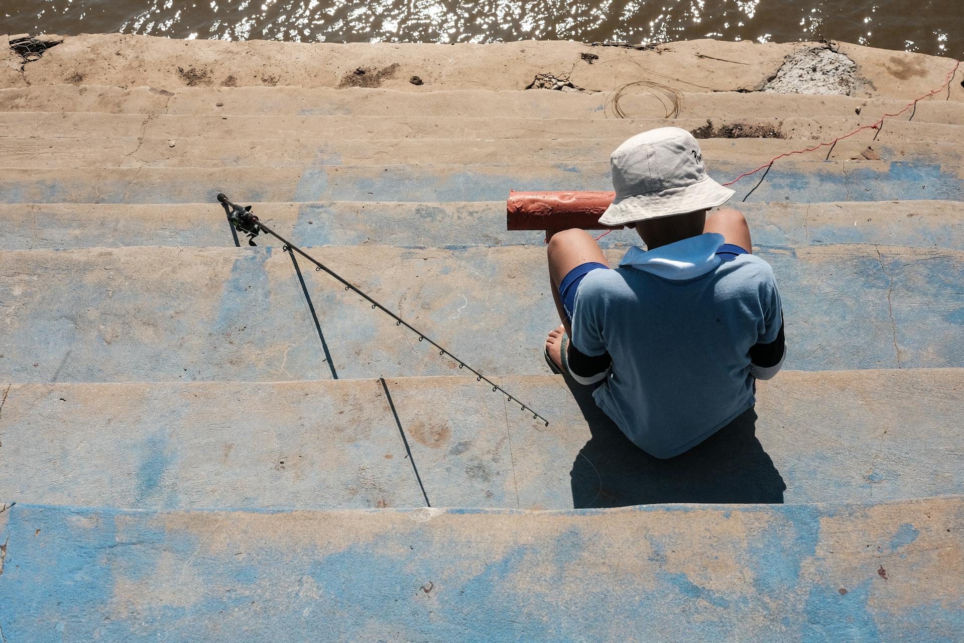 Man in a hat fishing on steps by a river in Asunción, Paraguay on a sunny day.