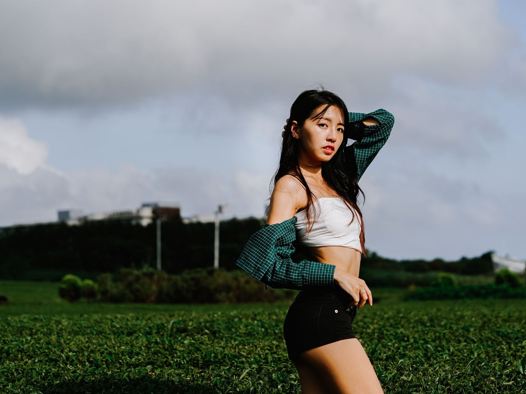 Fashionable woman posing outdoors in a lush green field under cloudy skies.