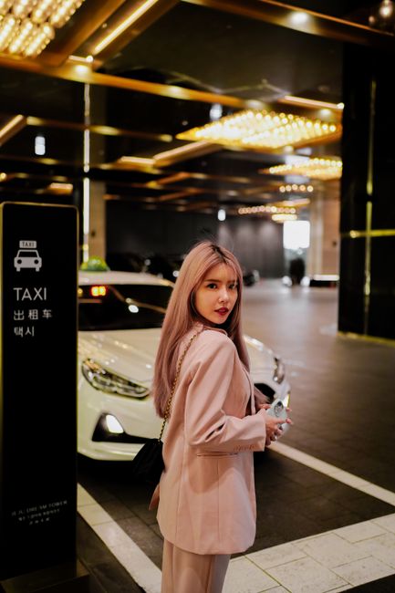 Fashionable woman with dyed hair stands in elegant outfit near taxi stand indoors.