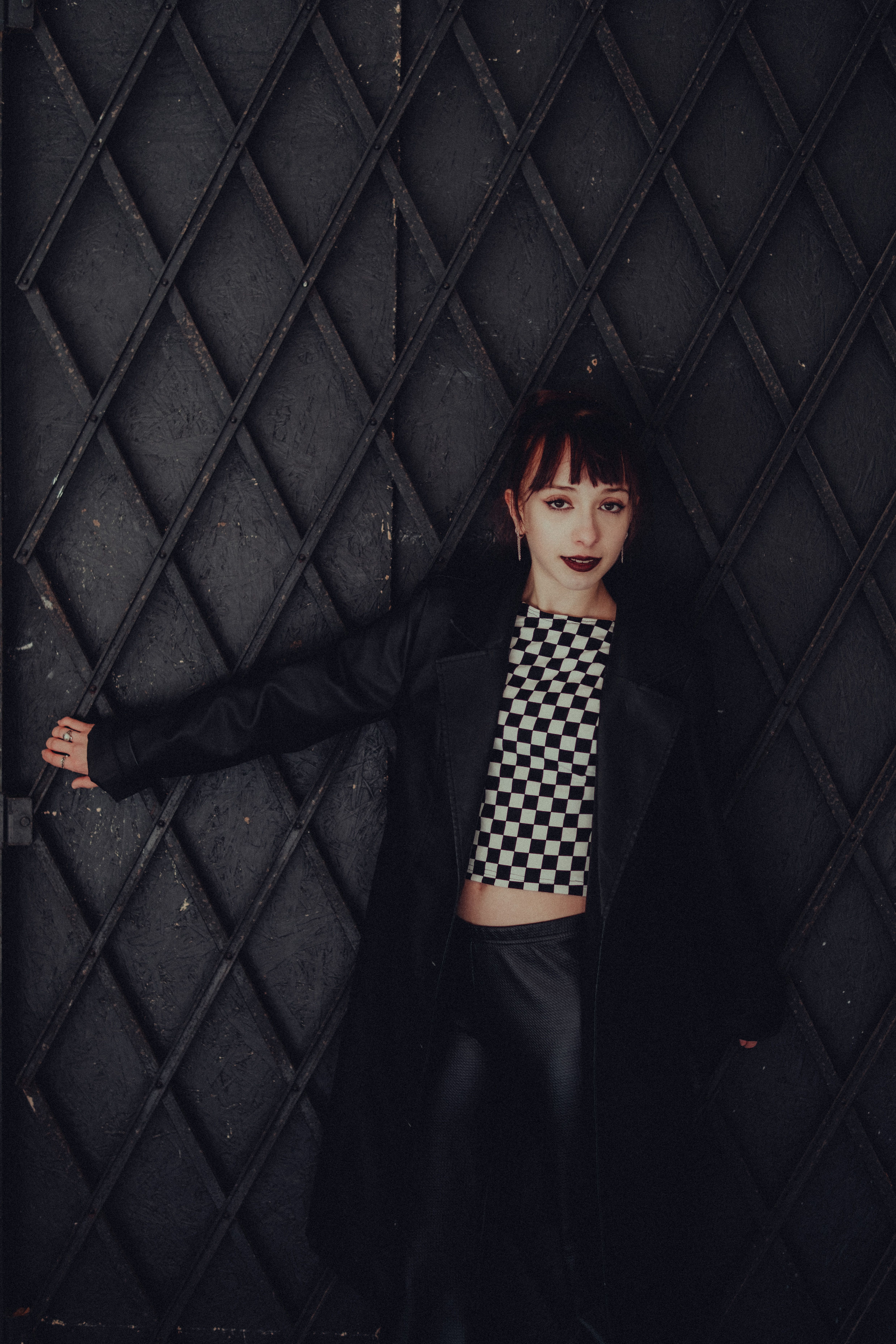A fashionable woman in a checkered top and jacket stands confidently against a textured wall.