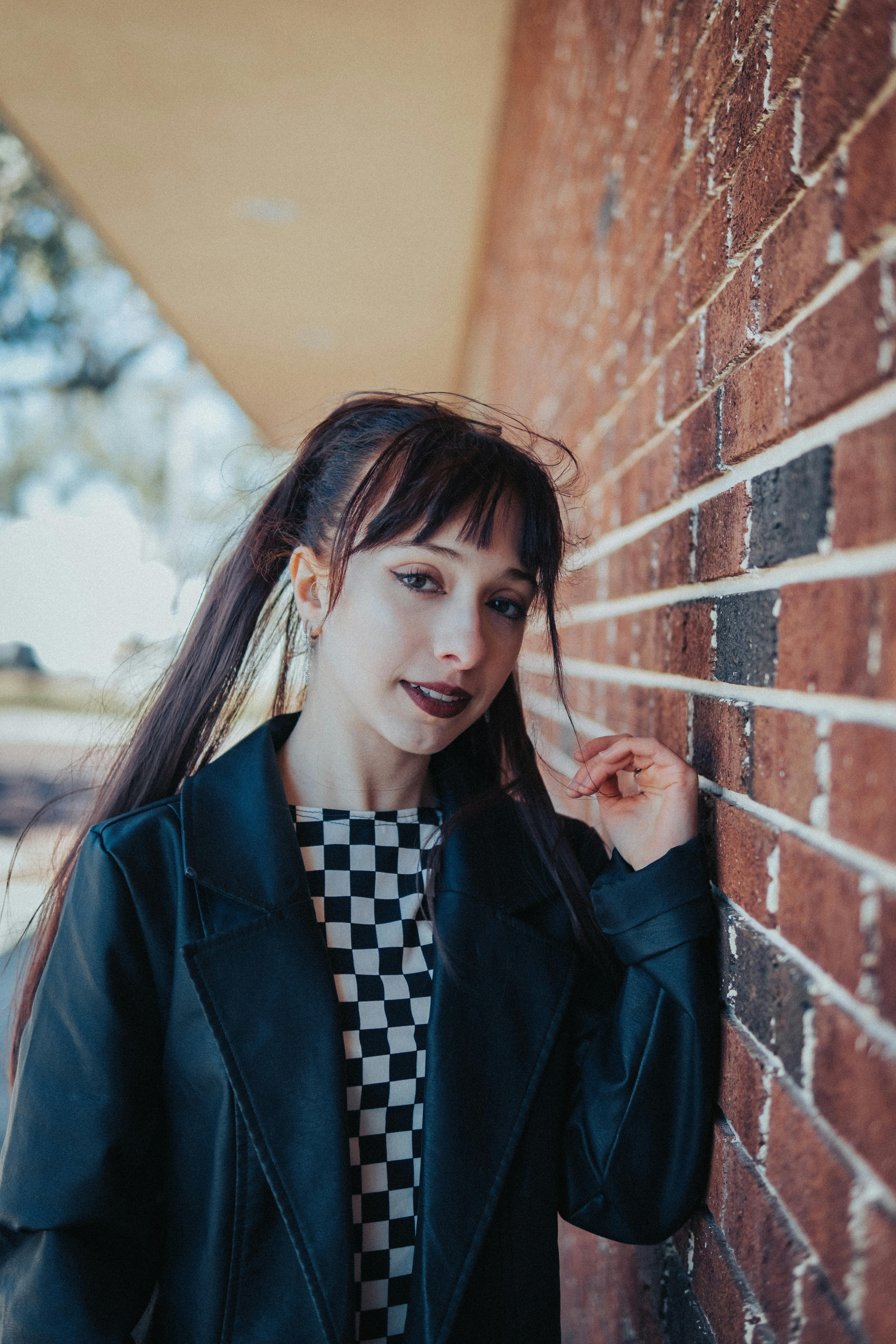 Stylish young woman with dark makeup posing outdoors against a brick wall on a sunny day.