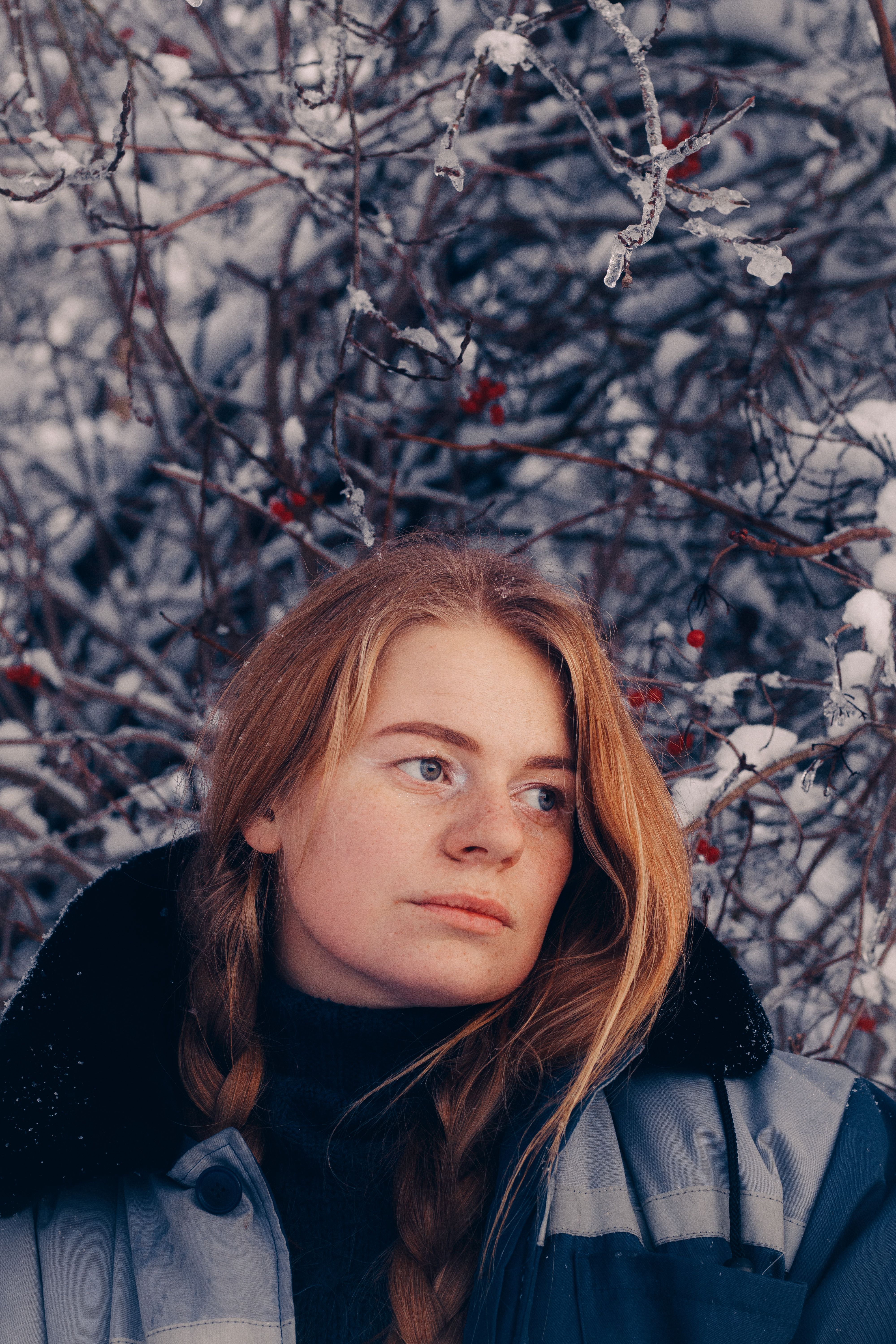 A young redhead woman with braided hair in a winter scene, surrounded by snow-covered branches.