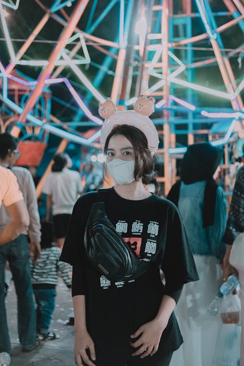 Girl Wearing a Face Mask Standing in an Amusement Park against a Ferris Wheel at Evening